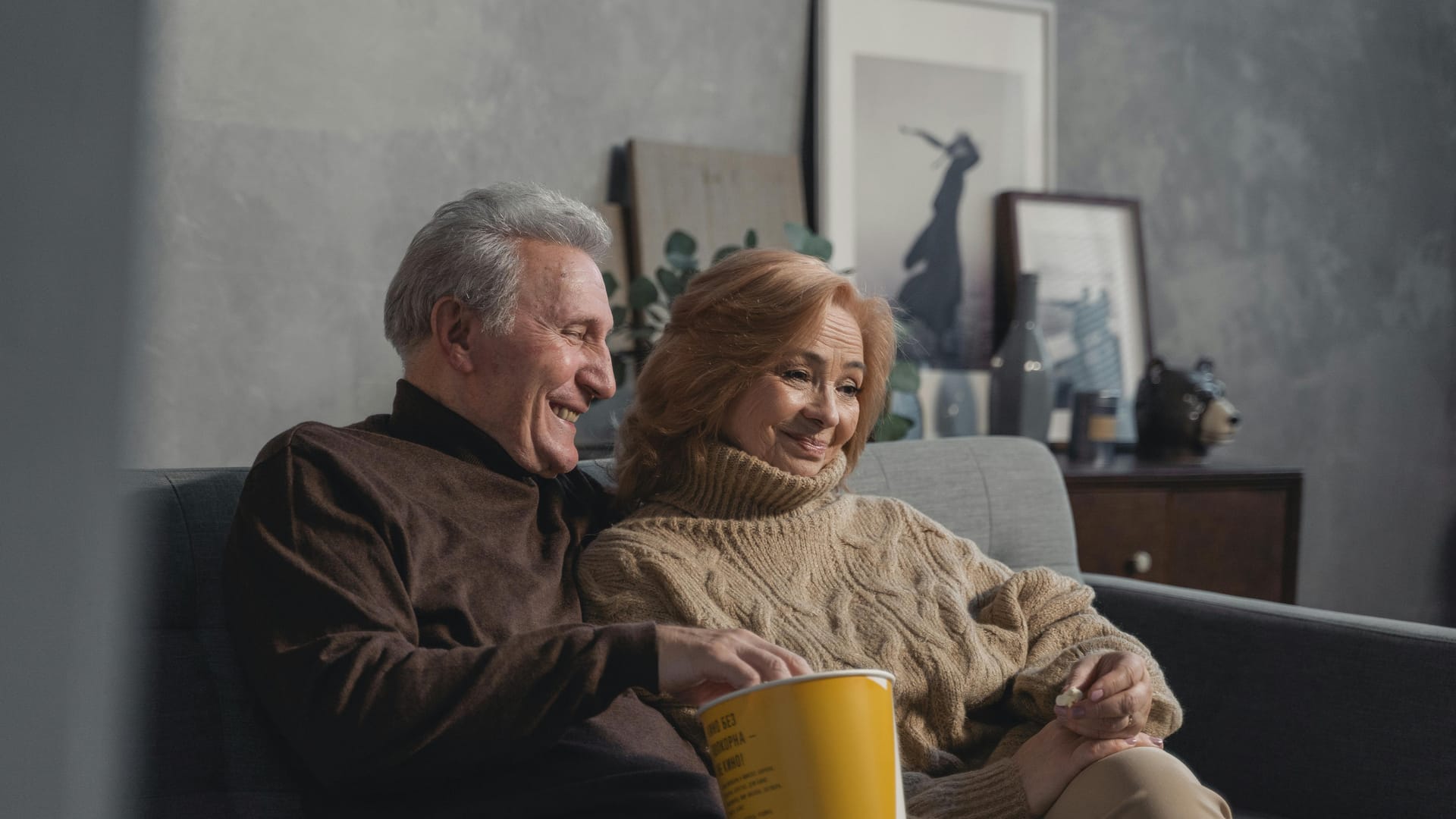 A senior couple laughing on a couch while sharing a bucket of popcorn