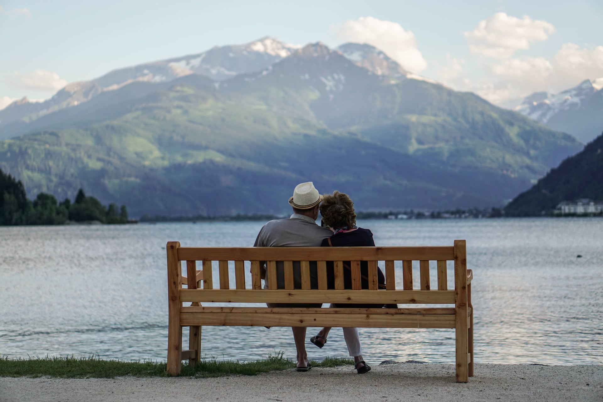 A senior couple on a wooden bench looking across a lake toward snow-capped mountains