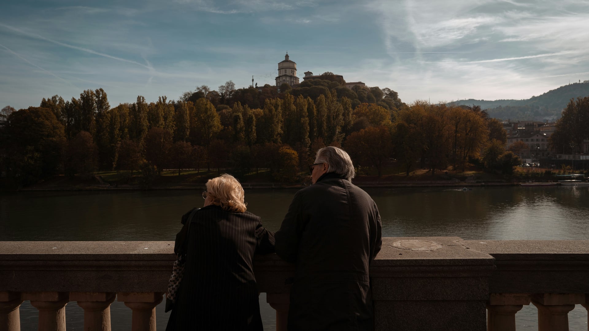 An older couple standing together at a river railing, looking out over the water