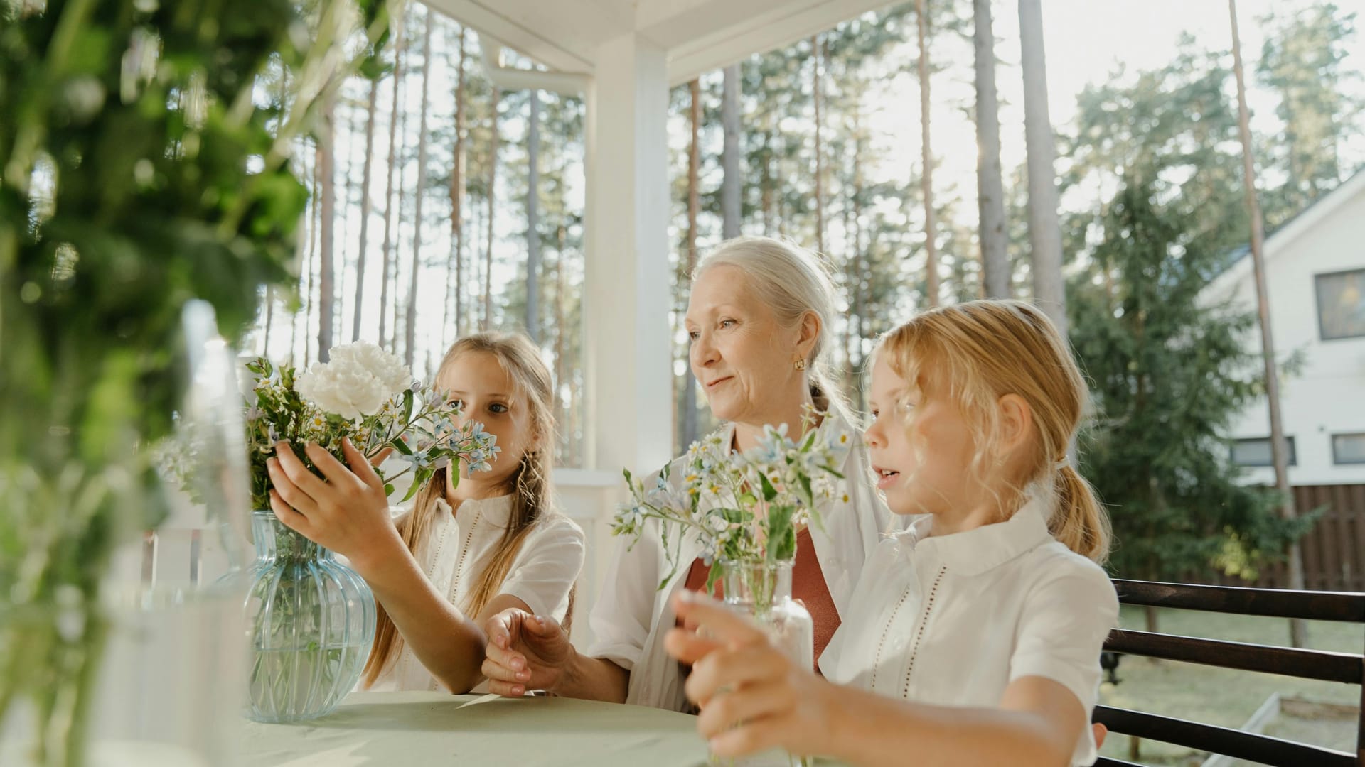 A grandmother arranging flowers with two young granddaughters on a sunlit porch